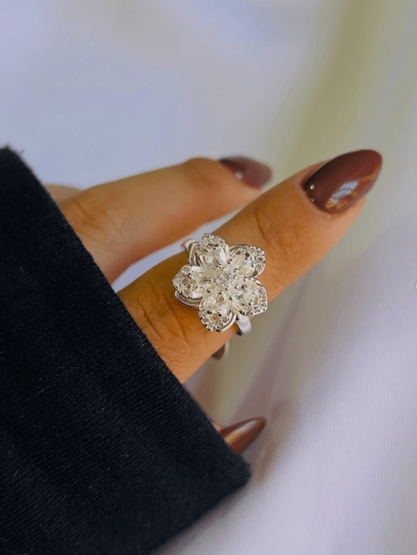 Mano con manicura marrón usando un anillo plateado grande con forma de flor y pedrería, sobre un fondo blanco