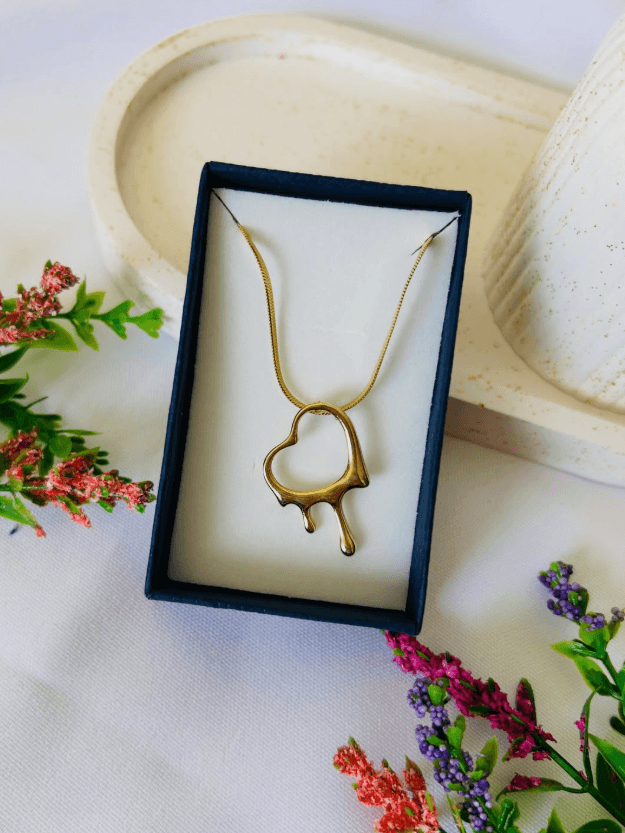 Collar dorado con un dije en forma de corazón que parece derretirse, presentado en una caja negra sobre una bandeja beige