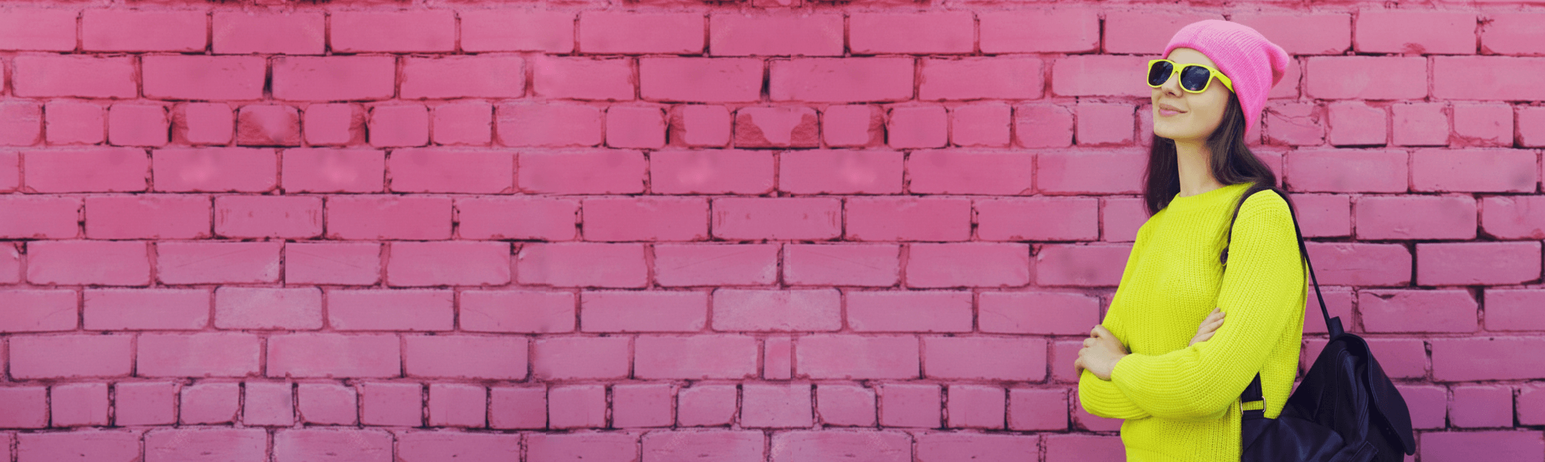 joven mujer sonriente con gorro rosa, gafas de sol y suéter verde neón, posando frente a una pared de ladrillo rosa 